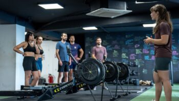 Séance de coaching HYROX en salle avec démonstration sur rameurs devant un groupe d’athlètes attentifs.