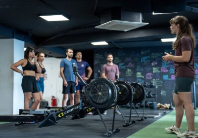 Séance de coaching HYROX en salle avec démonstration sur rameurs devant un groupe d’athlètes attentifs.