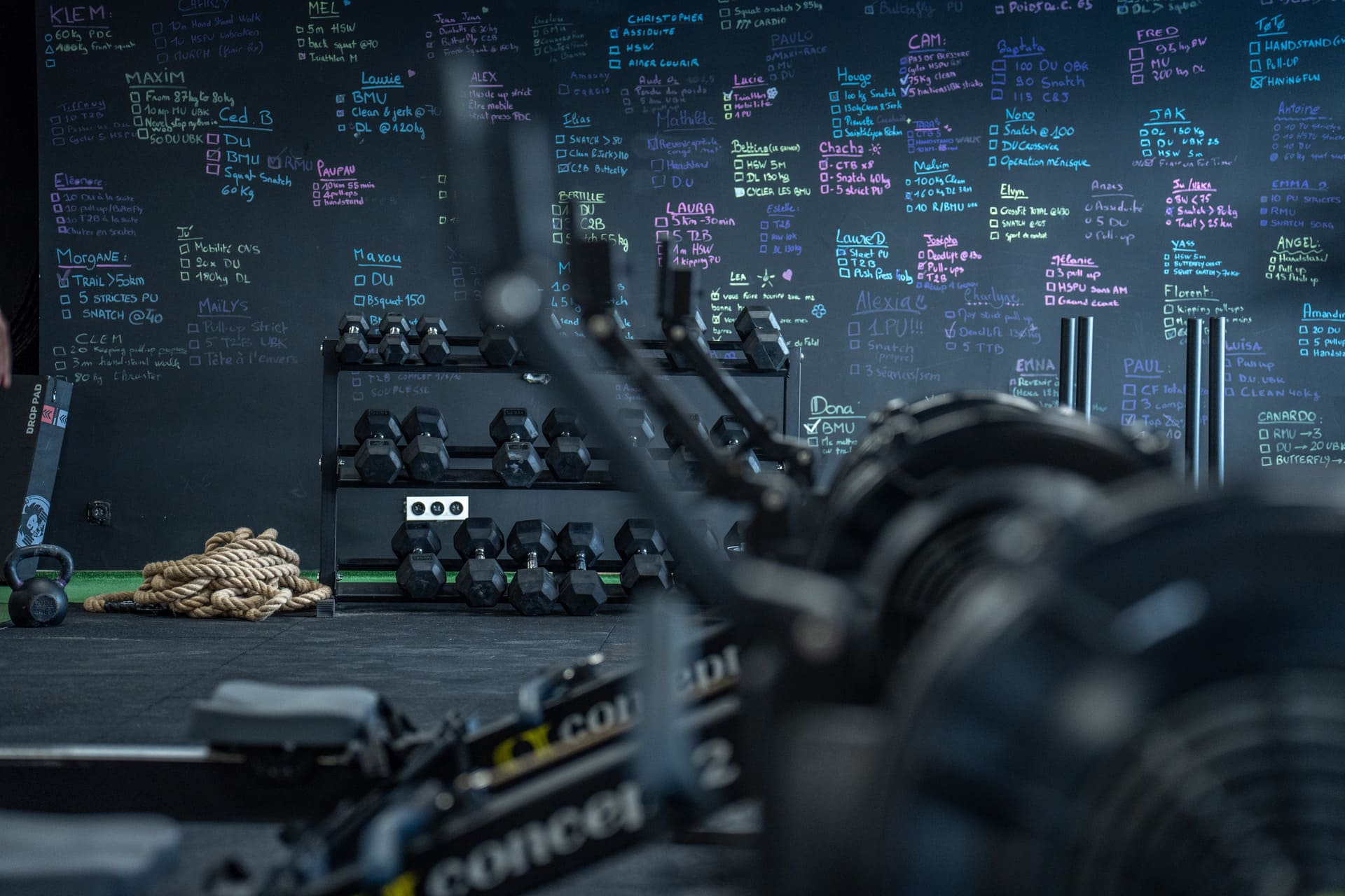 Salle d'entraînement HYROX avec rameurs, haltères, cordes, et tableau mural listant les performances des athlètes.